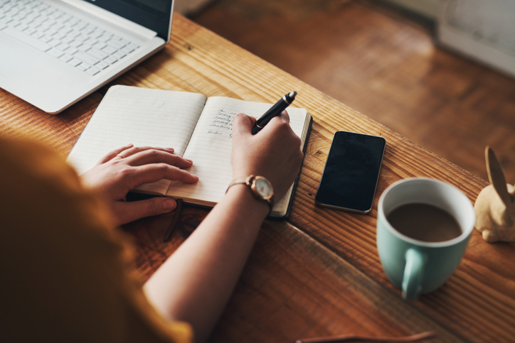 Shot of a woman's hand writing new year's resolutions in a notebook on a desk with a cup of coffee